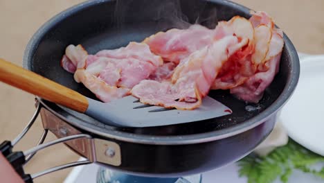 bacon slices begin to brown as spatula flips them inside smoky hot pan