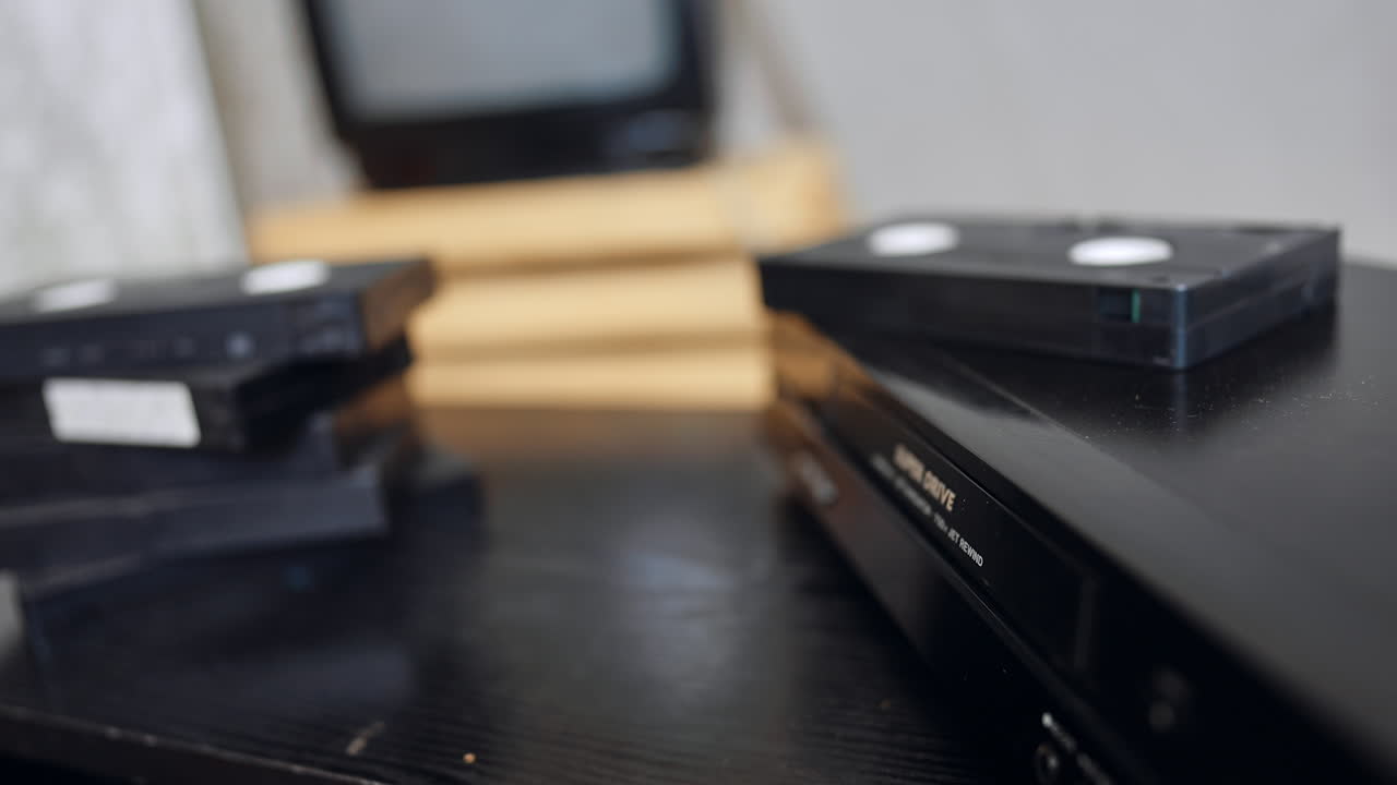 Senior male's hand puts the tape into VHS recorder. Close up. A pile of tapes and an old screen at blurred backdrop