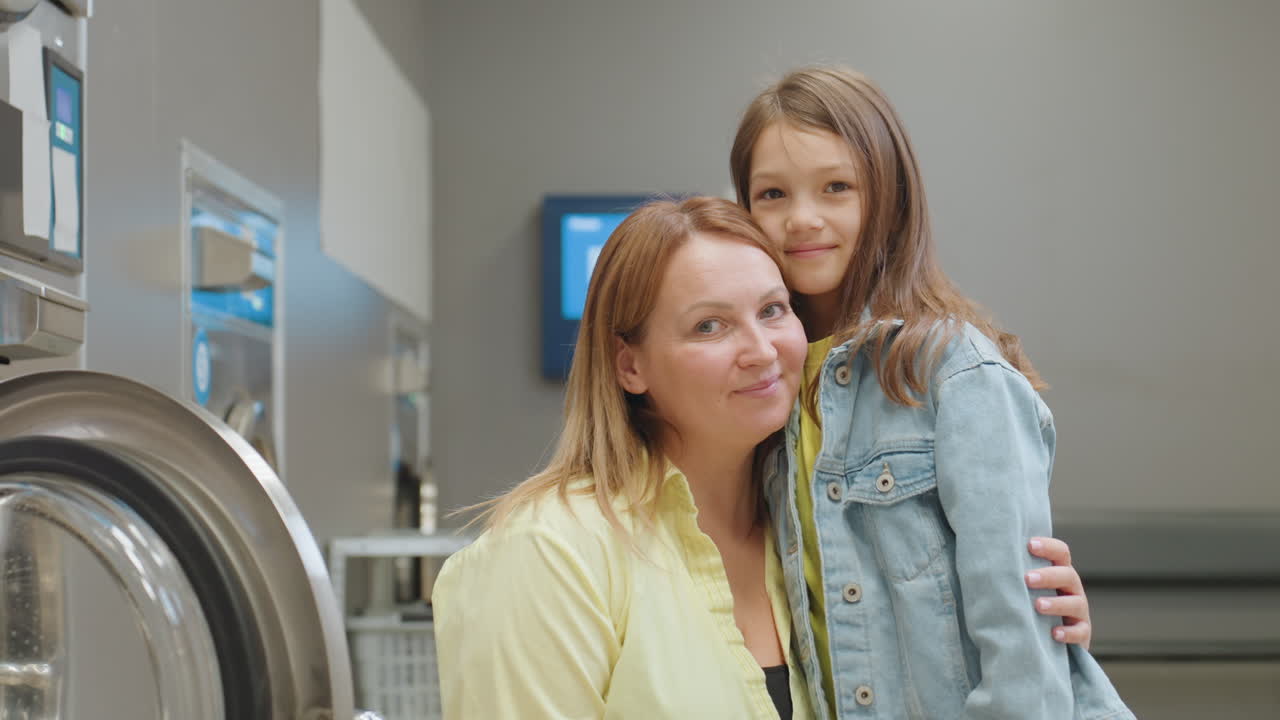 Mom and beautiful daughter share warm moment inside laundromat, smiling near washers, blurred payment machine in background, child in denim jacket, gentle hug, caring connection indoors