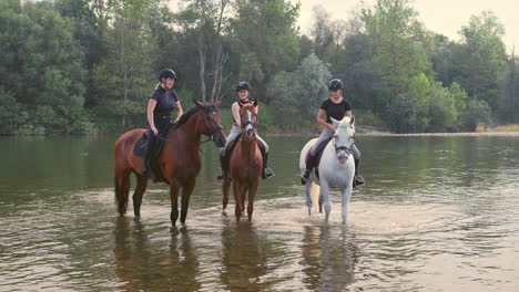 three women enjoying equestrian leisure and river, sitting on horses, wide shot