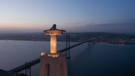 Aerial-wide-orbit-of-large-tall-monument-of-Santuario-de-Cristo-Rei-with-lights-and-red-Ponte-25-de-Abril-bridge-across-the-sea-near-the-coast-of-Lisbon,-Portugal-at-night