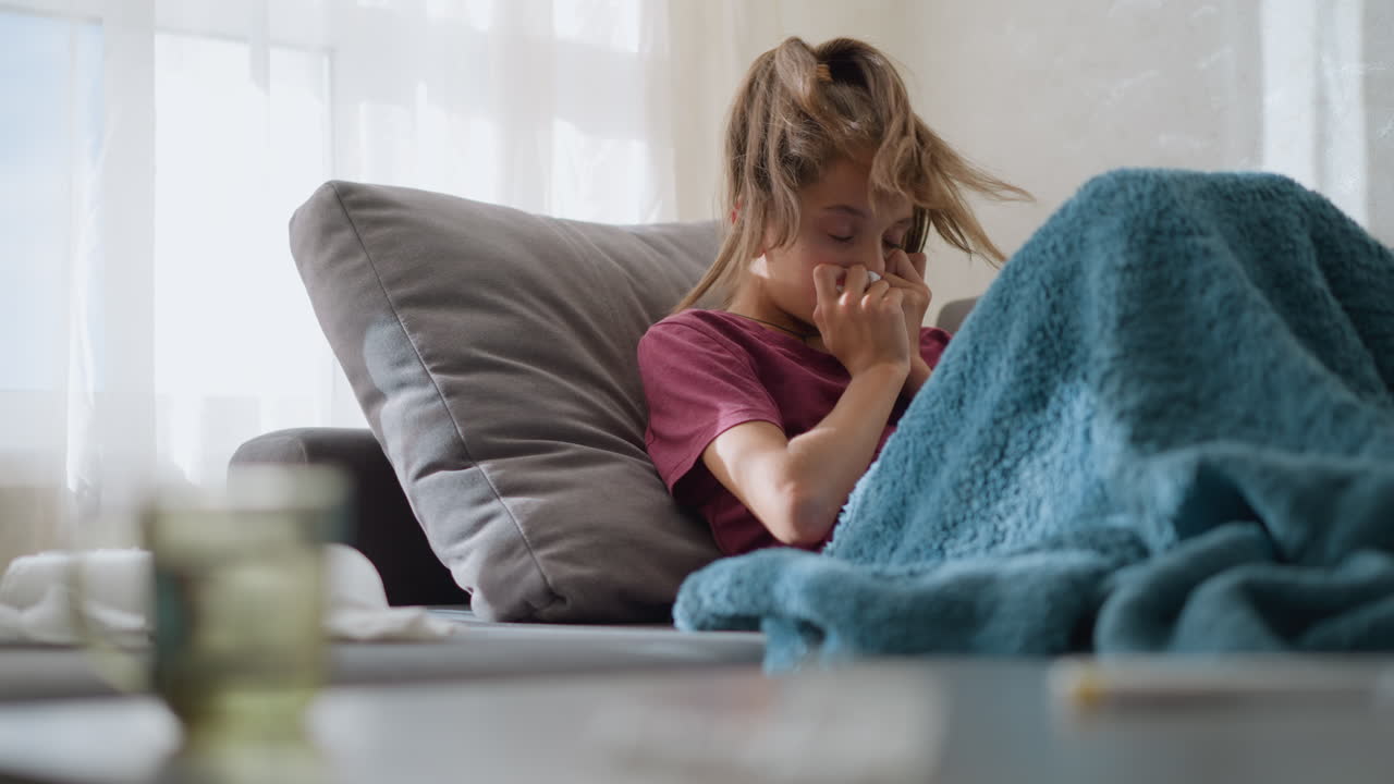 Primo piano sfocato di una tazza di vetro con un bambino sdraiato sul divano che soffre di influenza, riposa sotto una coperta, non si sente bene ed è a disagio a causa del raffreddore o dell'influenza, circondato da fazzoletti usati