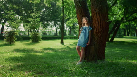 niña soñadora del tronco del árbol delgado en el parque. niño sonriente observando la naturaleza alrededor