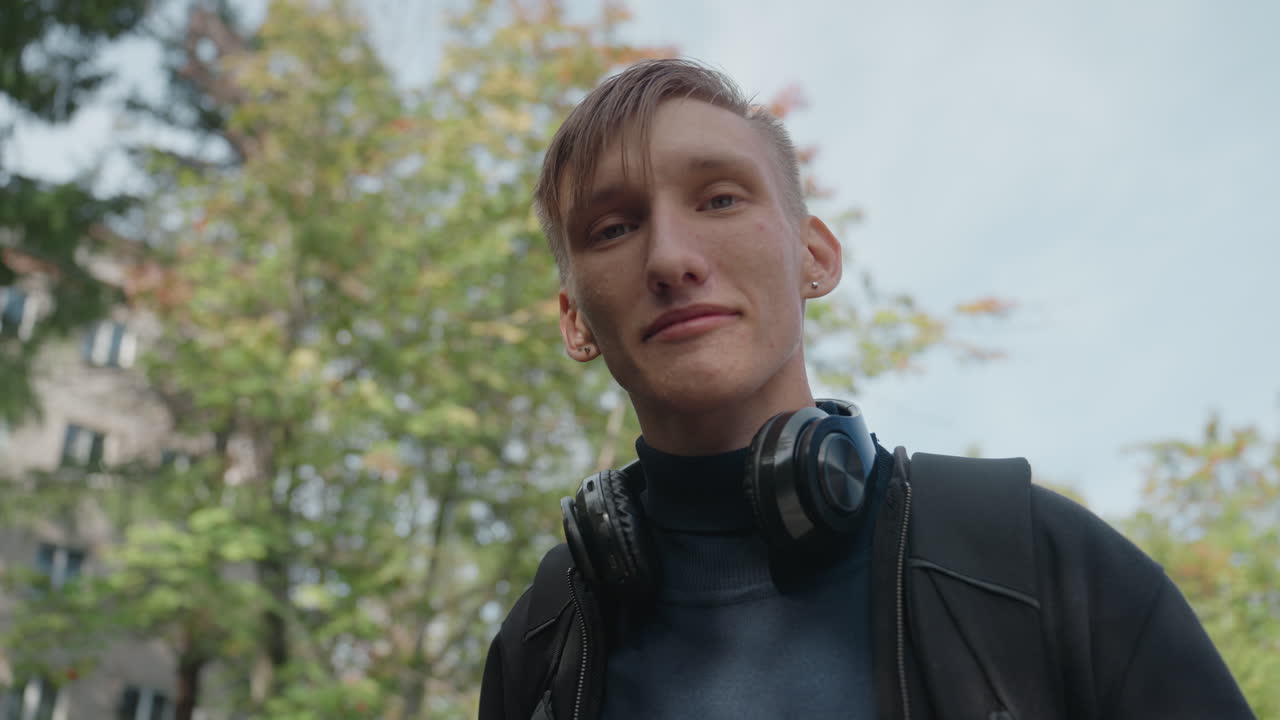 caucasian young man smiling with headphones closeup urban park portrait, warm expression, direct eye contact with camera, leafy backdrop, apartment facade, casual jacket, content creator vibe, upbeat