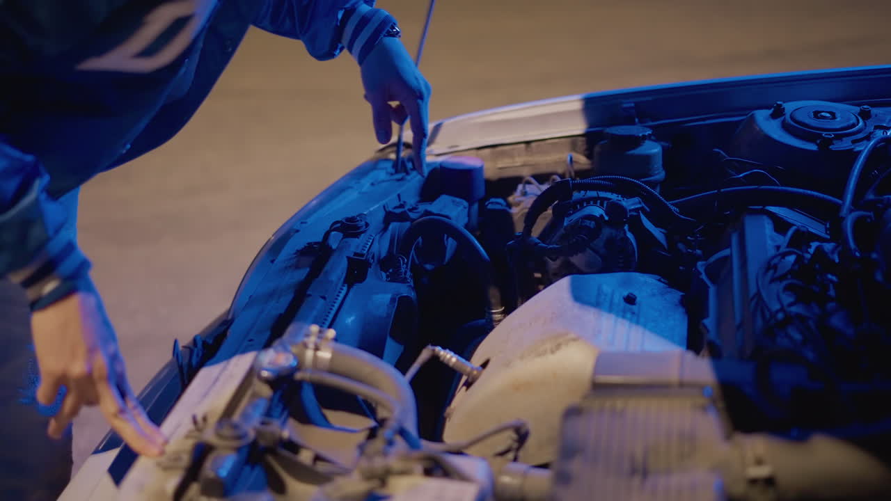 Coche averiado en una carretera de noche con el capó abierto. El conductor se inclina sobre el compartimento del motor bajo la luz azul, inspeccionando la batería y los cables. Ambiente tenso, primer plano de los movimientos de la mano, estacionamiento urbano vacío, capó levantado, solución rápida de problemas.