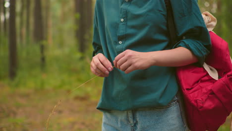 close-up of a woman walking along a forest trail, surrounded by tall trees, holding a strand of grass and playfully cutting it, while another woman with long nails is partially visible