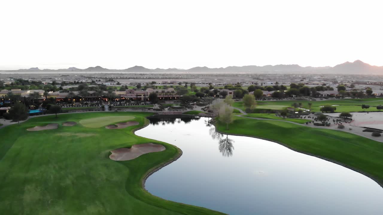 Drone footage from a golf course near a residential neighborhood in Arizona. High aerial shot of the greens, fairway, water harzard and clubhouse, with the Arizona mountains in the background