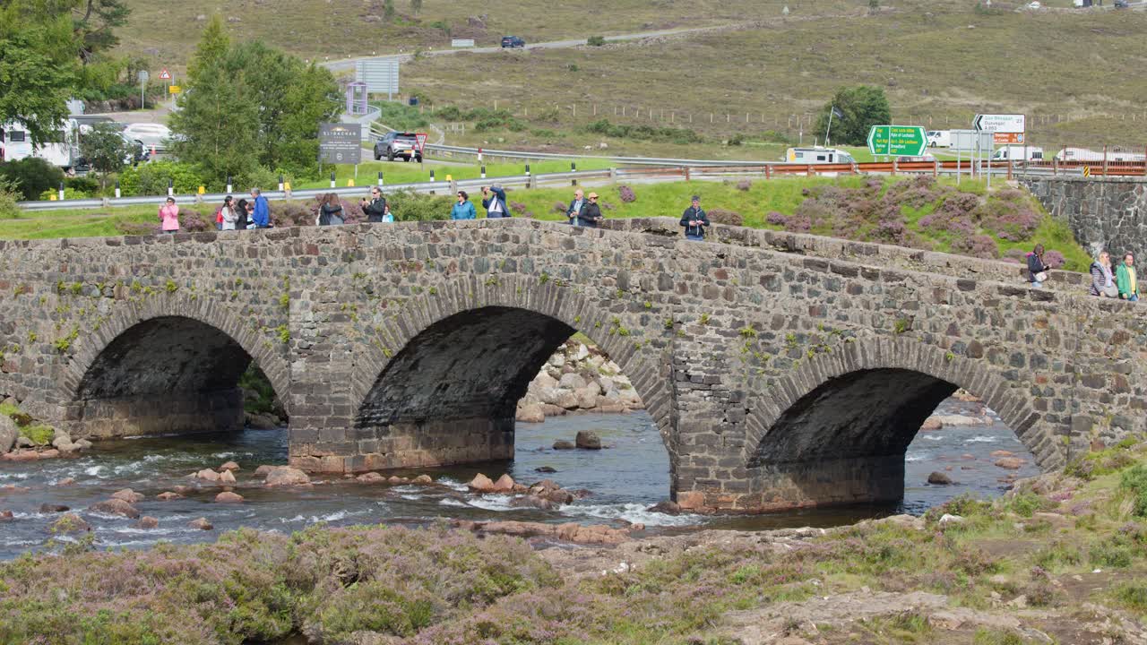 Tourists walk across arched stone bridge over river, daylight, wide shot, gentle camera pan