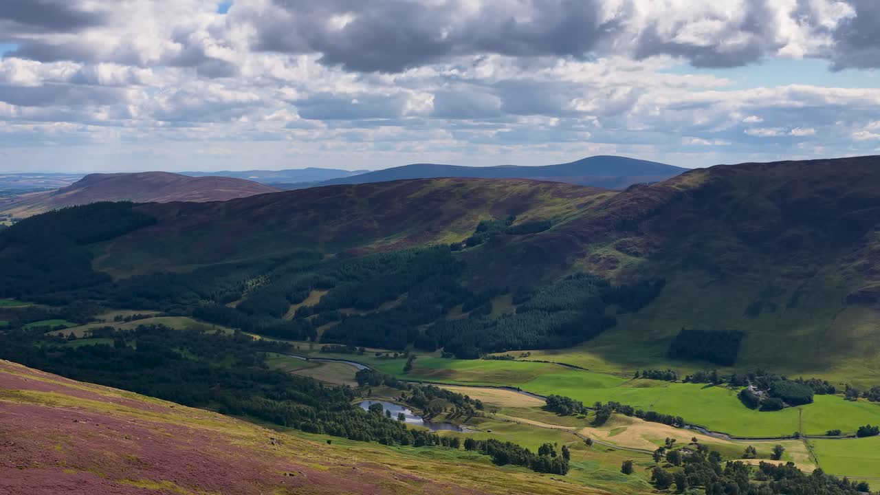 Aerial camera smoothly pans across sunlit Scottish Highlands valley, heather slopes, and dramatic cloudy sky