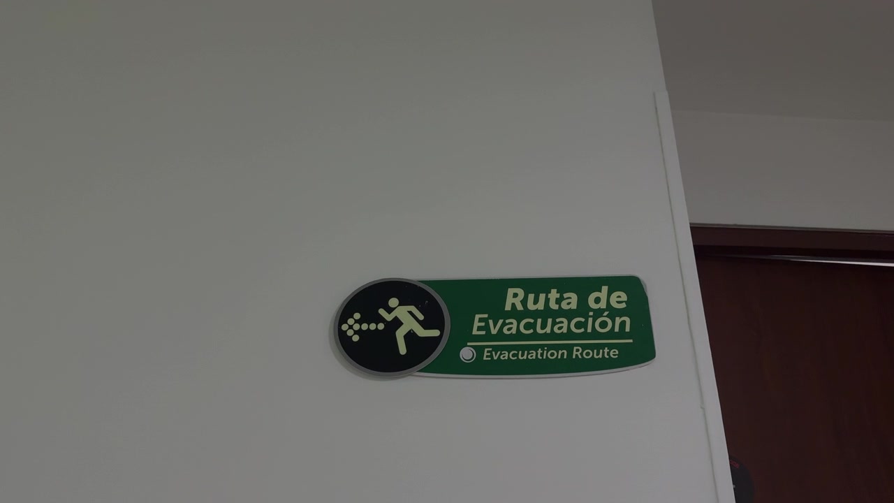 Groen tweetalig vluchtroutebord hangt aan het plafond in een witte gang terwijl de camera dichterbij komt onder tl-licht.