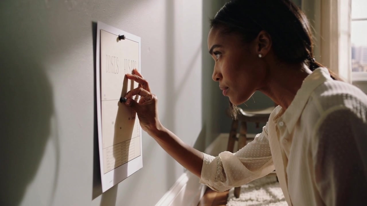 Donna in camicetta chiara che preme uno strumento per sfregare su carta appuntata, inginocchiata al muro, rivelando segni di sfregamento.