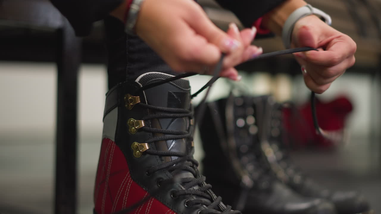 Closeup of lady tightening laces on red black ice skates with detailed view of hooks and fingers, preparation for skating indoors showing focus on equipment, hand movement and stylish polished nails