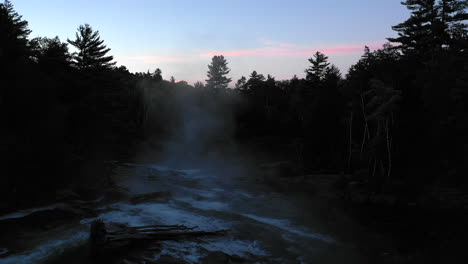 aerial drone shot over a dark misty pond and stream at sunset with forest trees in silhouette