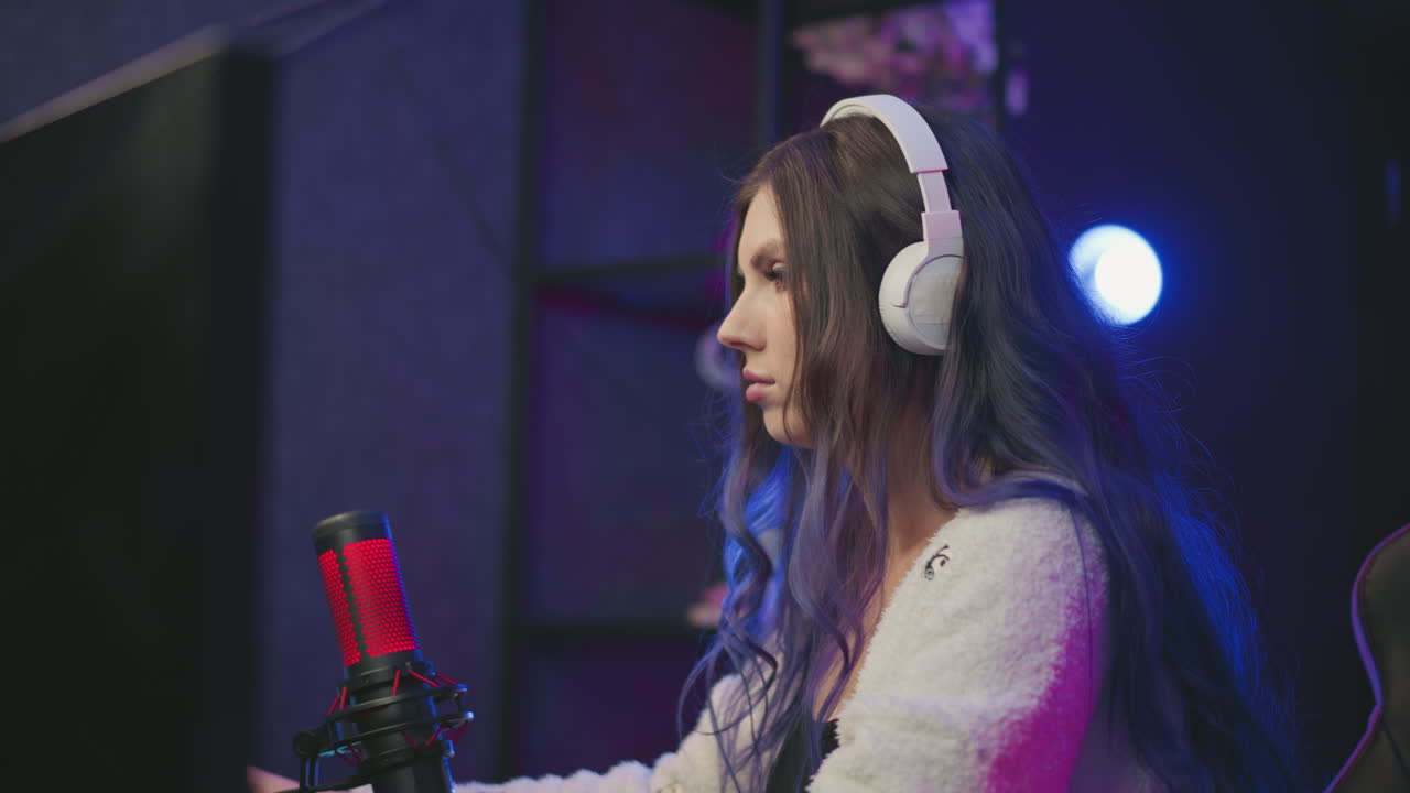 young woman wearing white headphones intently stares at monitor while playing game with condenser microphone in front of her, seated in gaming chair under moody ambient lighting in stylish setup