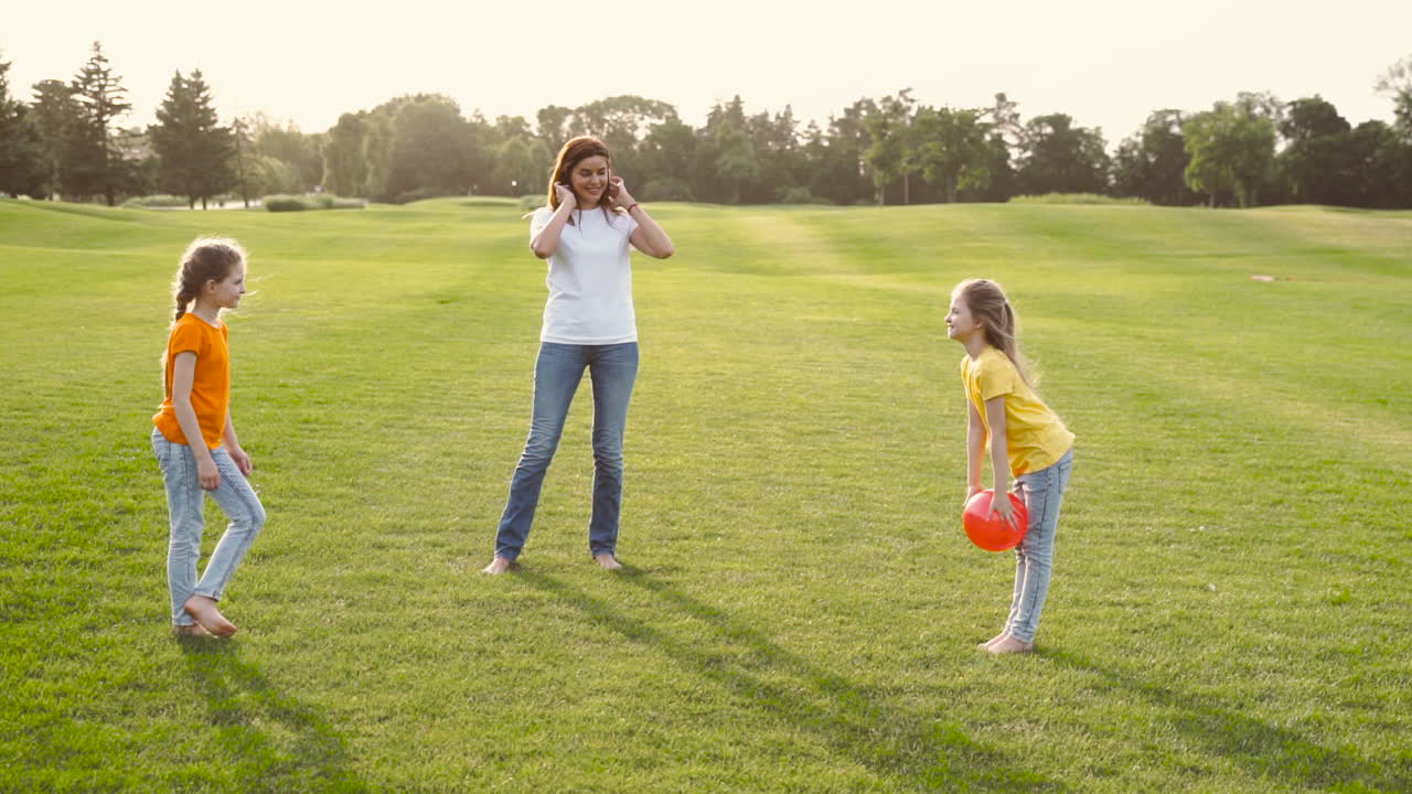 Happy Mother And Her Two Little Daughters Playing With Ball On Meadow