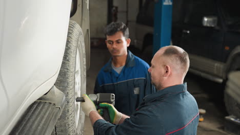 close-up of mechanic in blue uniform using a pneumatic tool to tighten wheel bolts of a lifted car in a workshop, while a colleague attentively observes the process, ensuring precision