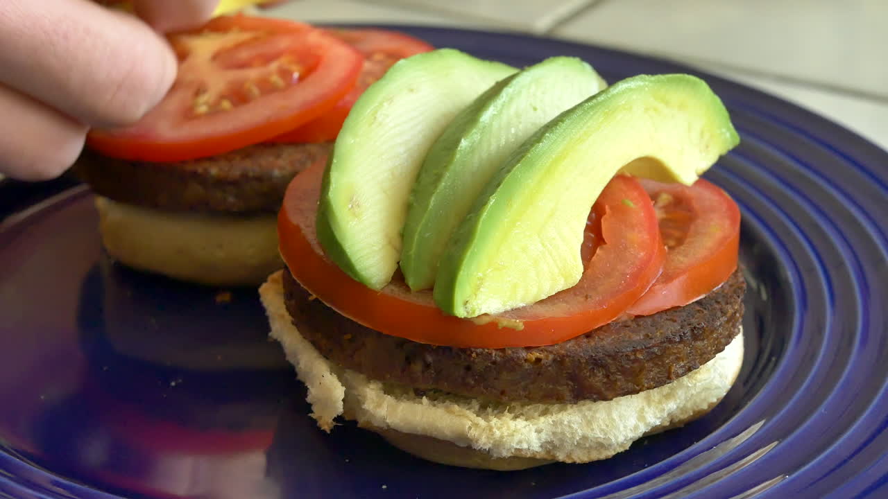 un chef vegano cocinando y ensamblando dos hamburguesas vegetarianas saludables de tomate y aguacate en un plato azul en la cocina