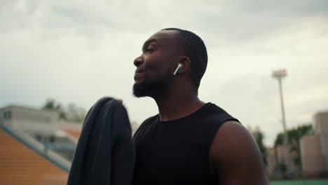 an athlete with black skin in a black t-shirt wipes his face with a towel and drinks water from a special bottle on a city beach against a gray sky