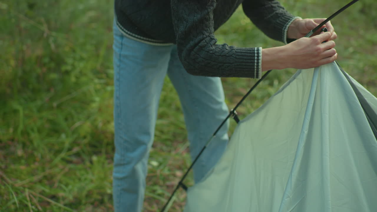 Tourist rises and secures tent fabric onto flexible pole during setup in forest as sunlight softly glows on surroundings, highlighting effort in peaceful nature camping environment