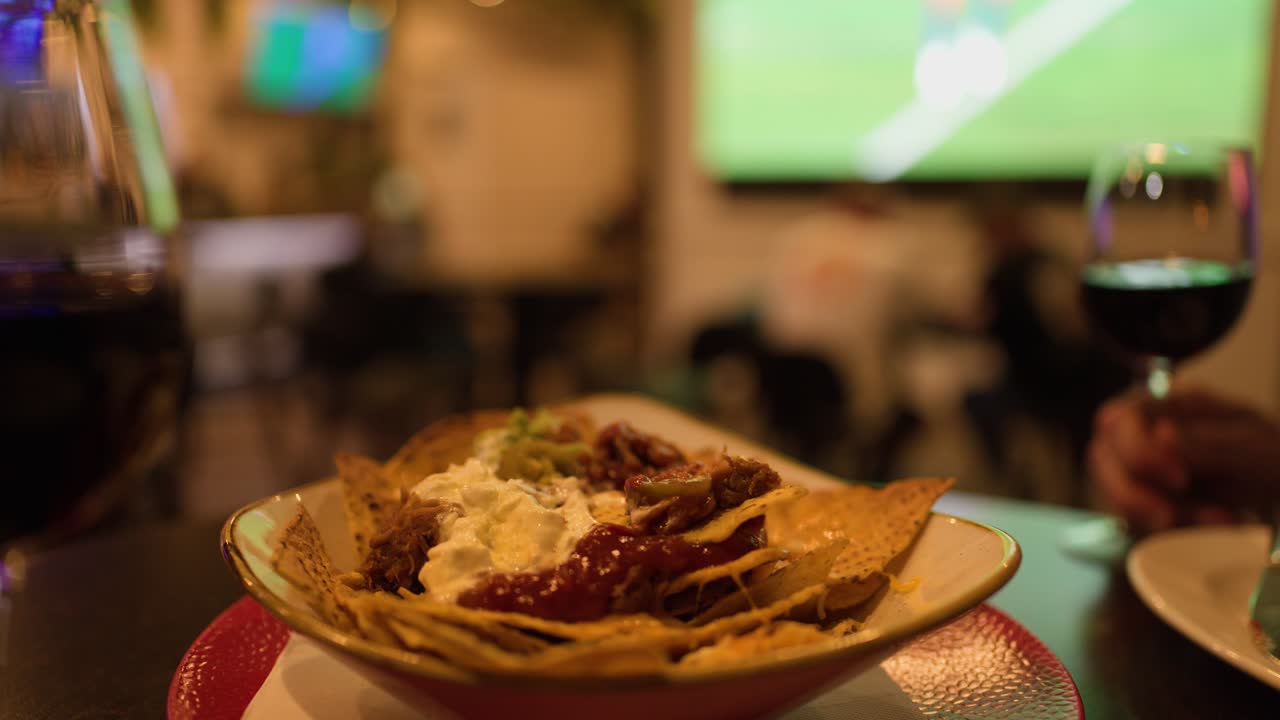 Hand reaches for nachos with salsa, sour cream, wine, sports game on TV, warm lighting