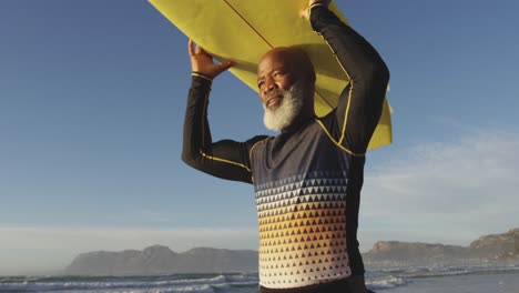 hombre afroamericano mayor caminando con una tabla de surf en la cabeza en la playa