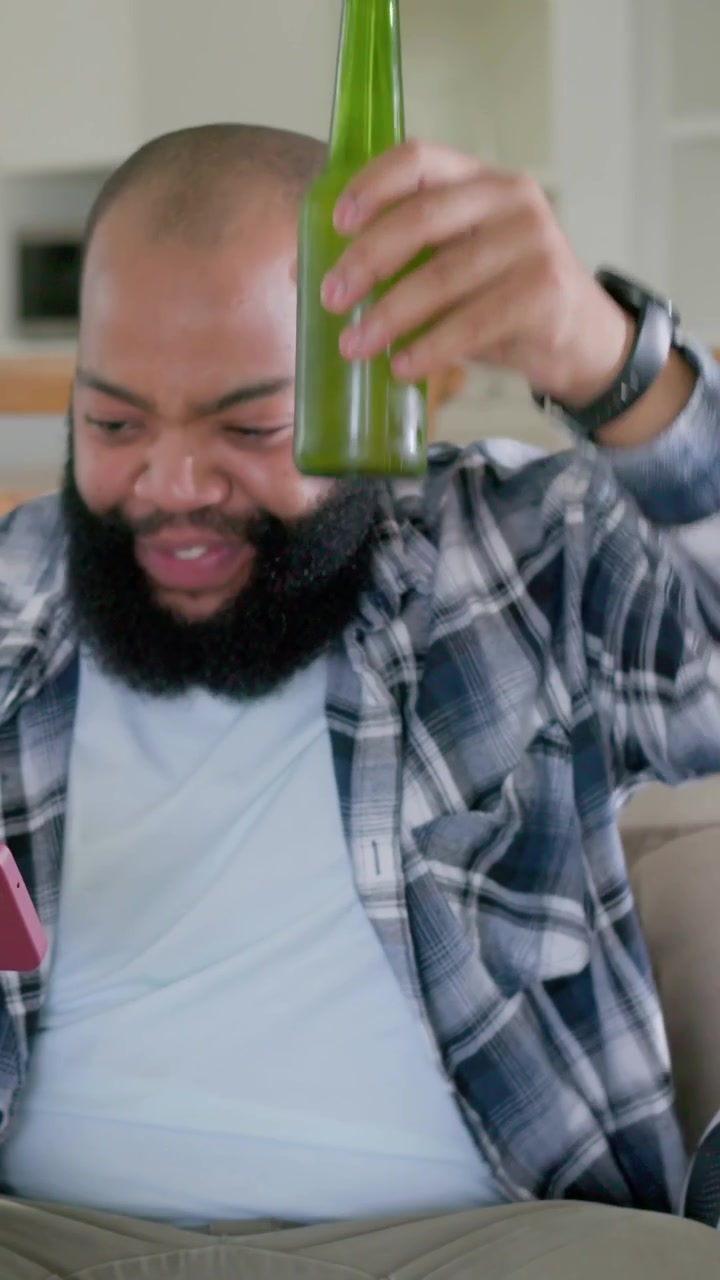 Vertical video: Reacting to red phone content, man sipping green bottle on couch in living room