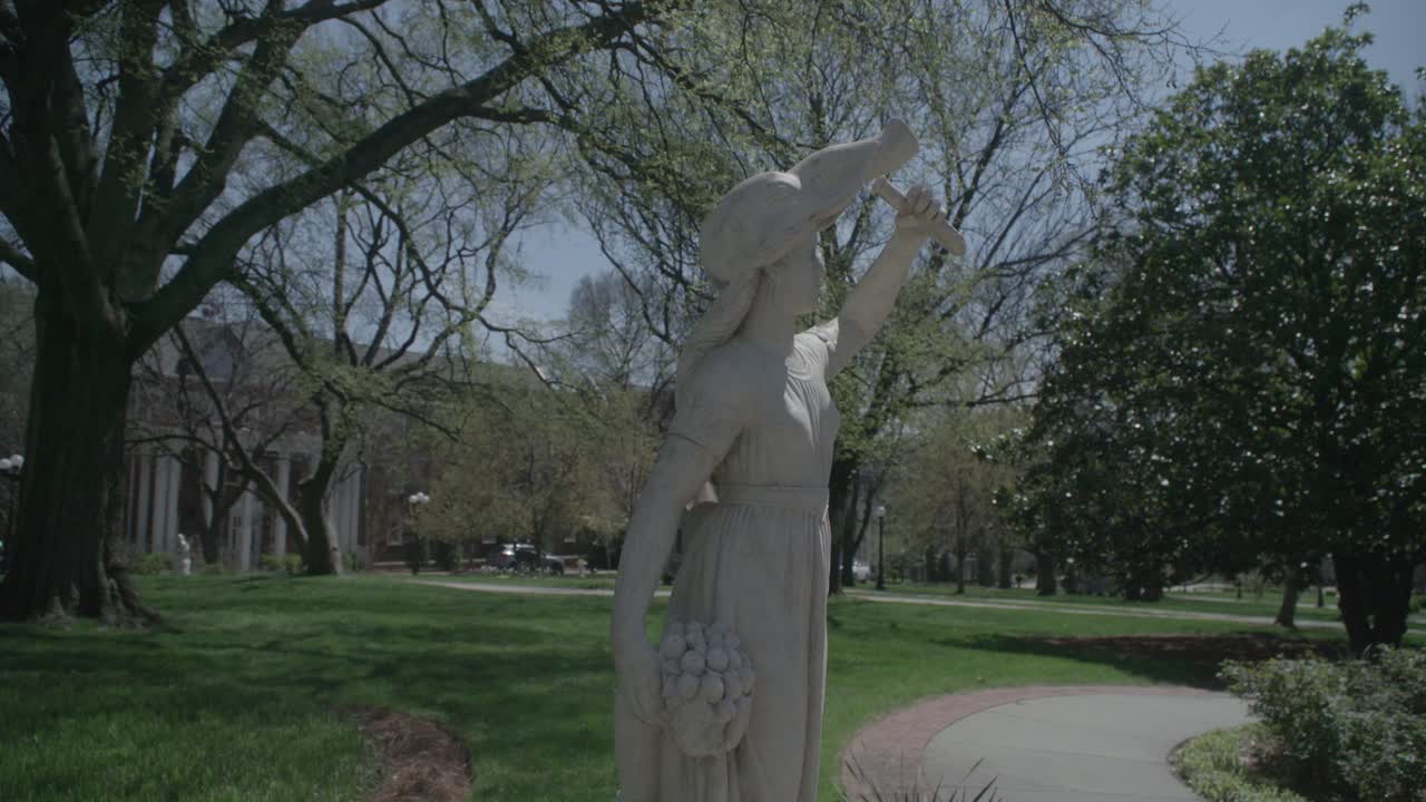 una toma media de una estatua clásica de una mujer en un jardín, sus brazos levantados con gracia, enmarcada por árboles frondosos, con arquitectura tradicional en el fondo en nashville
