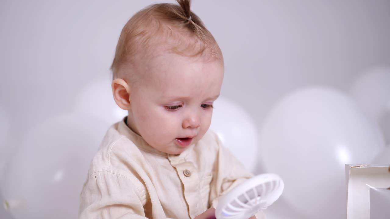 Adorable Caucasian baby with tiny tail on his head holding a small fan. Cute child smiles sweetly to camera. Close up.