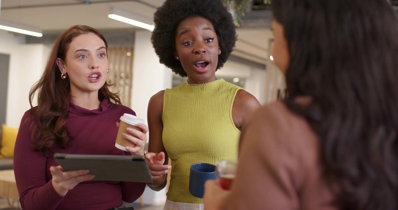 Diverse female coworkers approaching in office lounge, maroon-top tapping tablet, reviewing deck