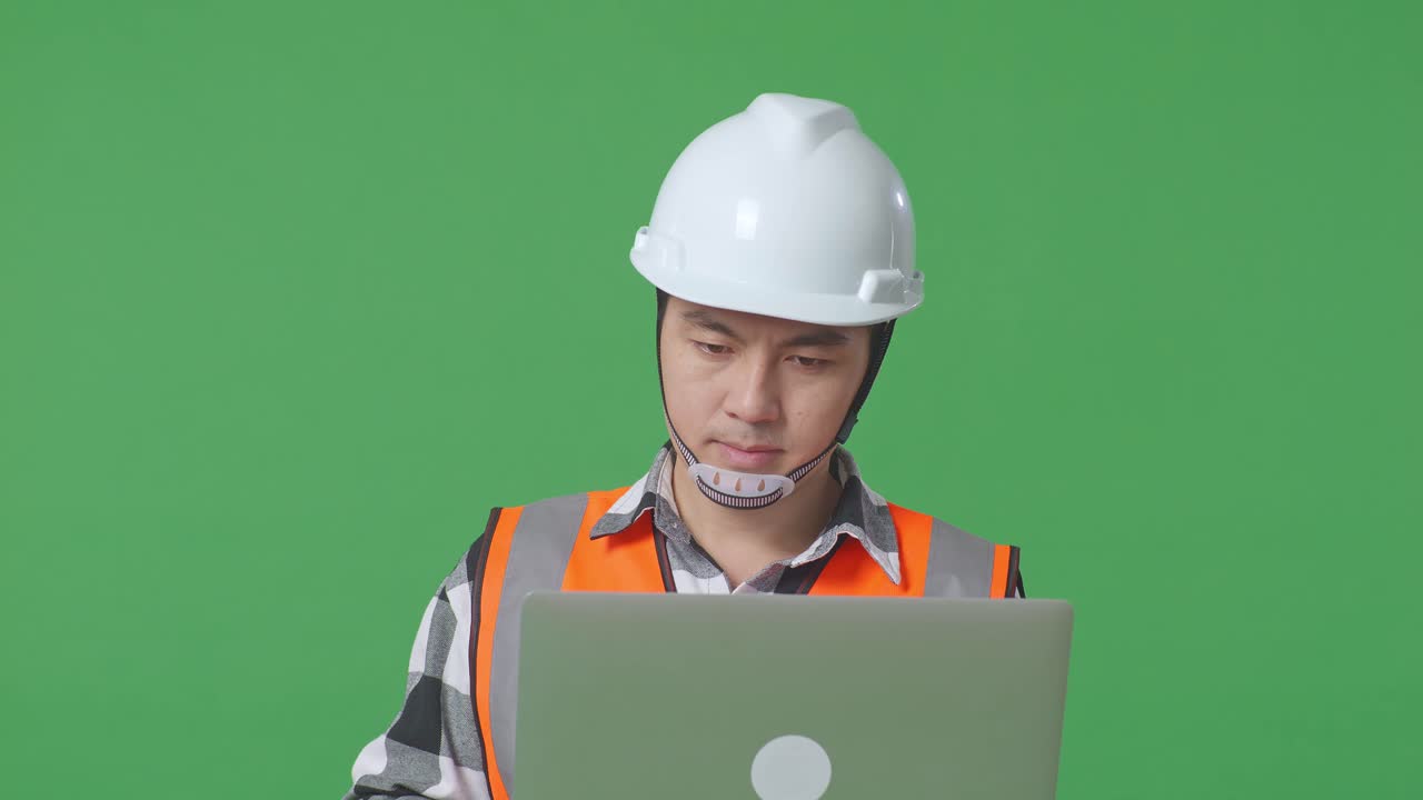 Close Up Of Asian Male Engineer With Safety Helmet Working On A Laptop And Looking Around While Standing In The Green Screen Background Studio