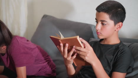 cute kid reading novel on couch with focused expression while sister beside him concentrates on her own task showing peaceful study environment and sibling focus in quiet shared learning moment