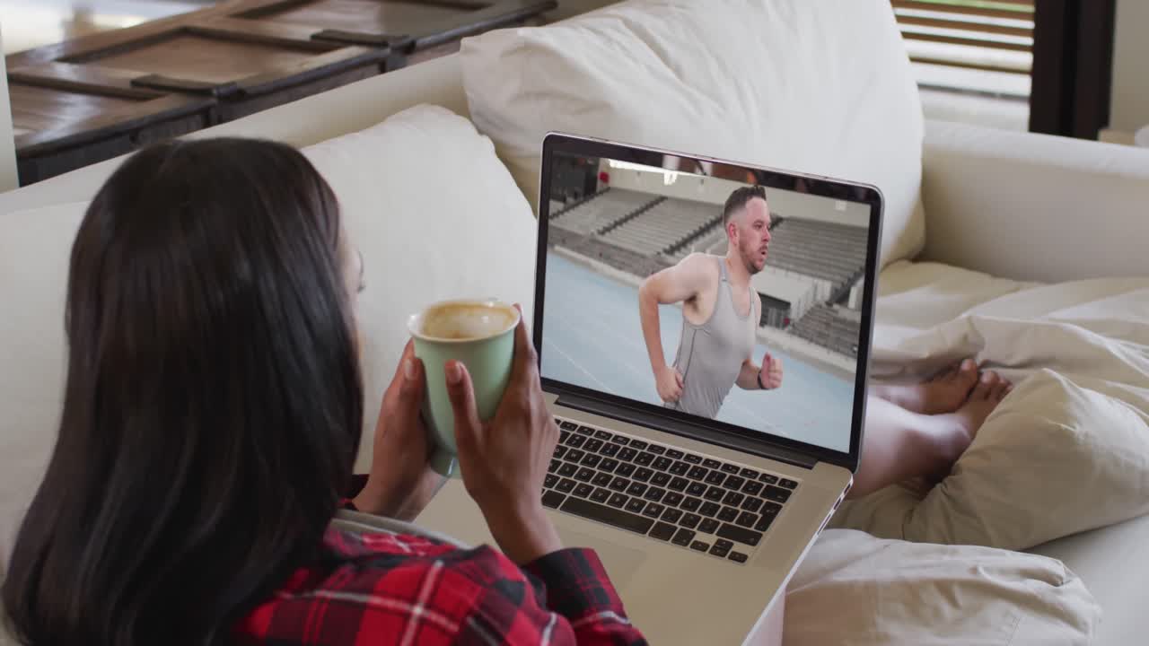 Composite of woman sitting at home holding coffee watching athletics event on laptop