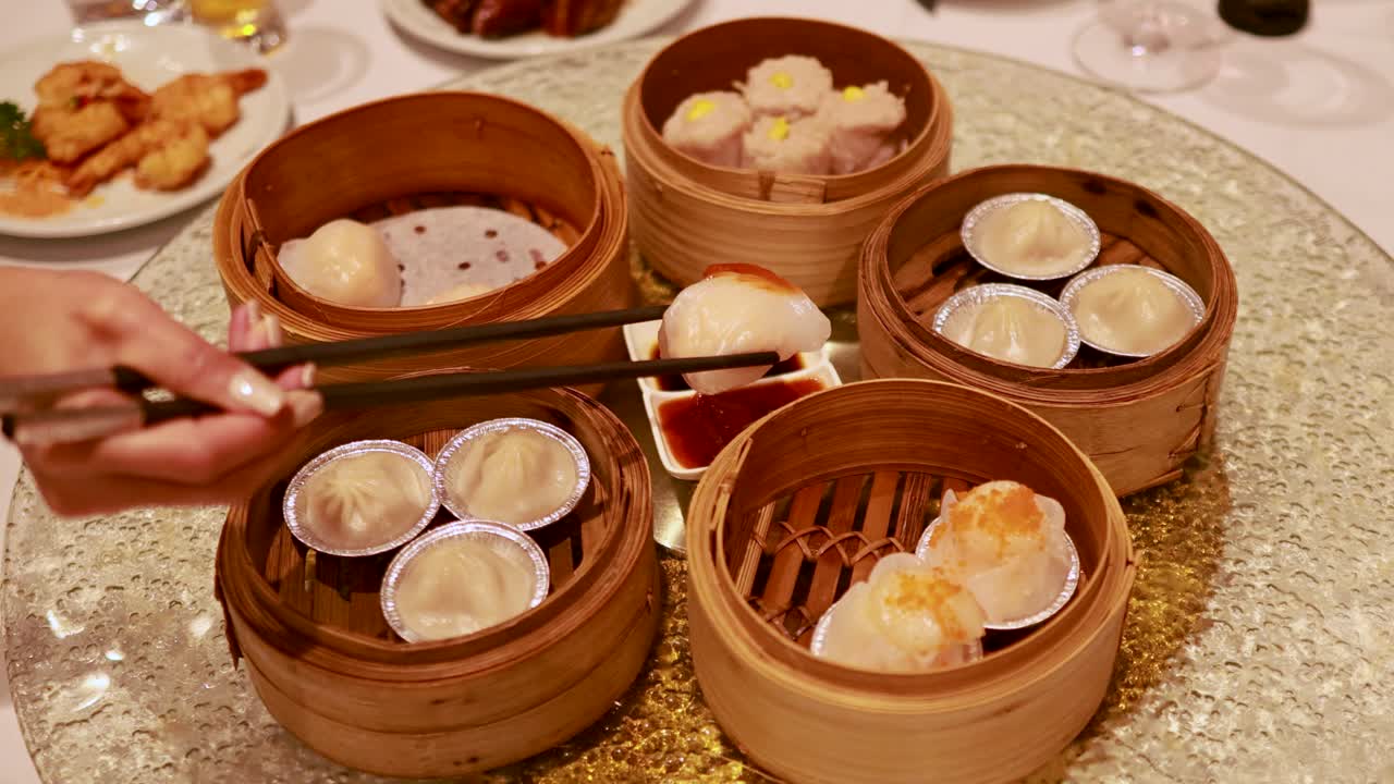 Hands use chopsticks to pick dim sum from bamboo steamers on a rotating table, creating a communal dining experience