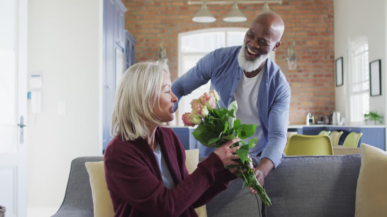 Glückliches, diverses Seniorenpaar sitzt im Wohnzimmer auf dem Sofa, überreicht Blumen und lächelt.