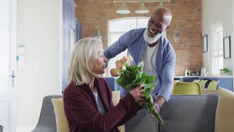 feliz pareja diversa en la sala de estar sentada en el sofá, dando flores y sonriendo