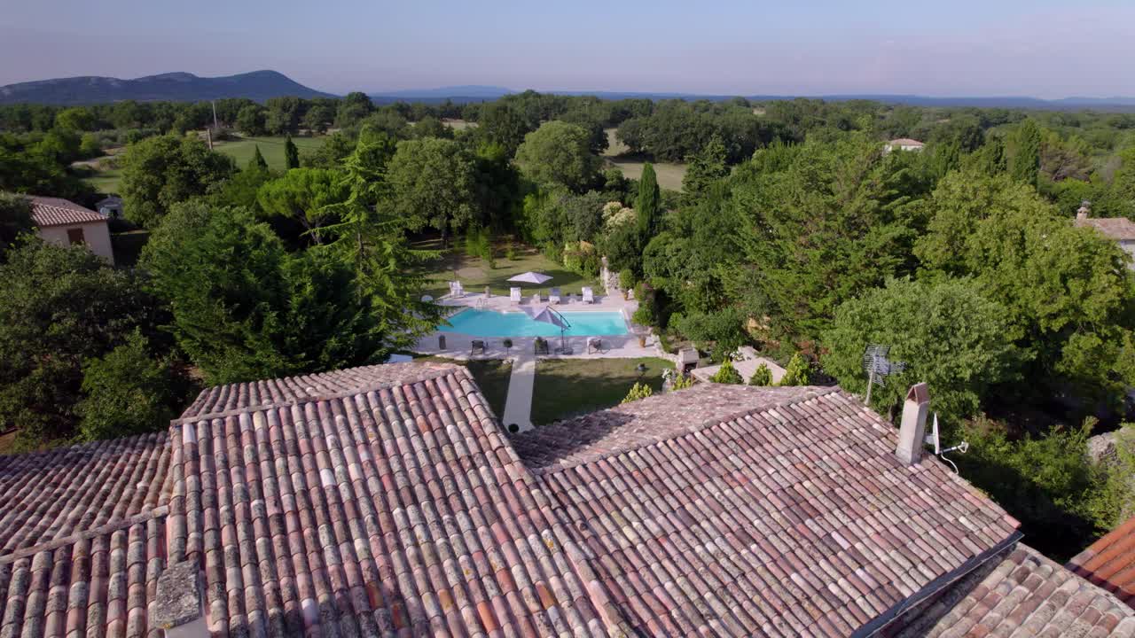 aerial view taken by drone of the courtyard of a stone villa in the south of france with a light-colored tiled roof, a garden revealed in the background with a beautiful swimming pool, sunny weather.