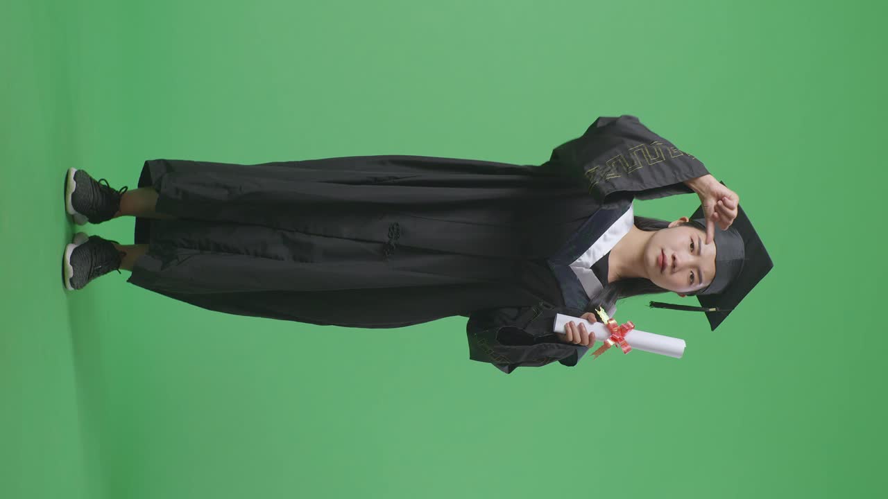 Studentessa asiatica laureata a figura intera con tocco, toga e diploma, che indica la telecamera e mostra il pollice verso il basso su sfondo green screen in studio