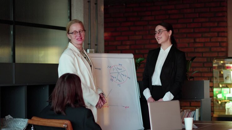 A confident bespectacled blonde businesswoman in a white business suit presents her project together with her colleague, a brunette girl in glasses and a black business suit during a meeting in the office