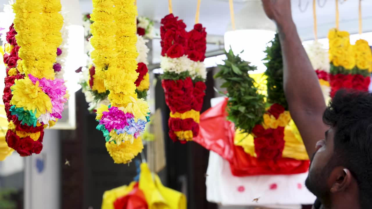 Un uomo sistema vivaci ghirlande di fiori in una bancarella di un mercato all'aperto a Little India, Singapore, sotto la luce del giorno brillante con un'inquadratura stabile della telecamera.