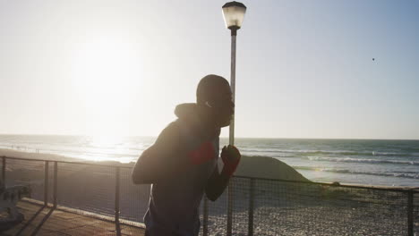 Hombre-Afroamericano-Enfocado-Boxeando-Y-Corriendo,-Haciendo-Ejercicio-Al-Aire-Libre-Junto-Al-Mar