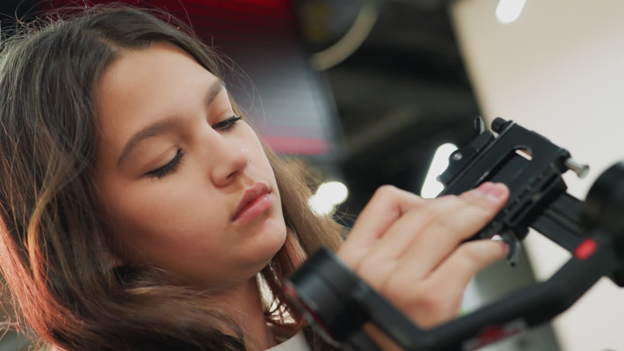 teen student adjusting camera gimbal concentrating on setup, indoor studio environment, hands mounting lens and stabilizer, testing movement and frame, learning practical filmmaking skills