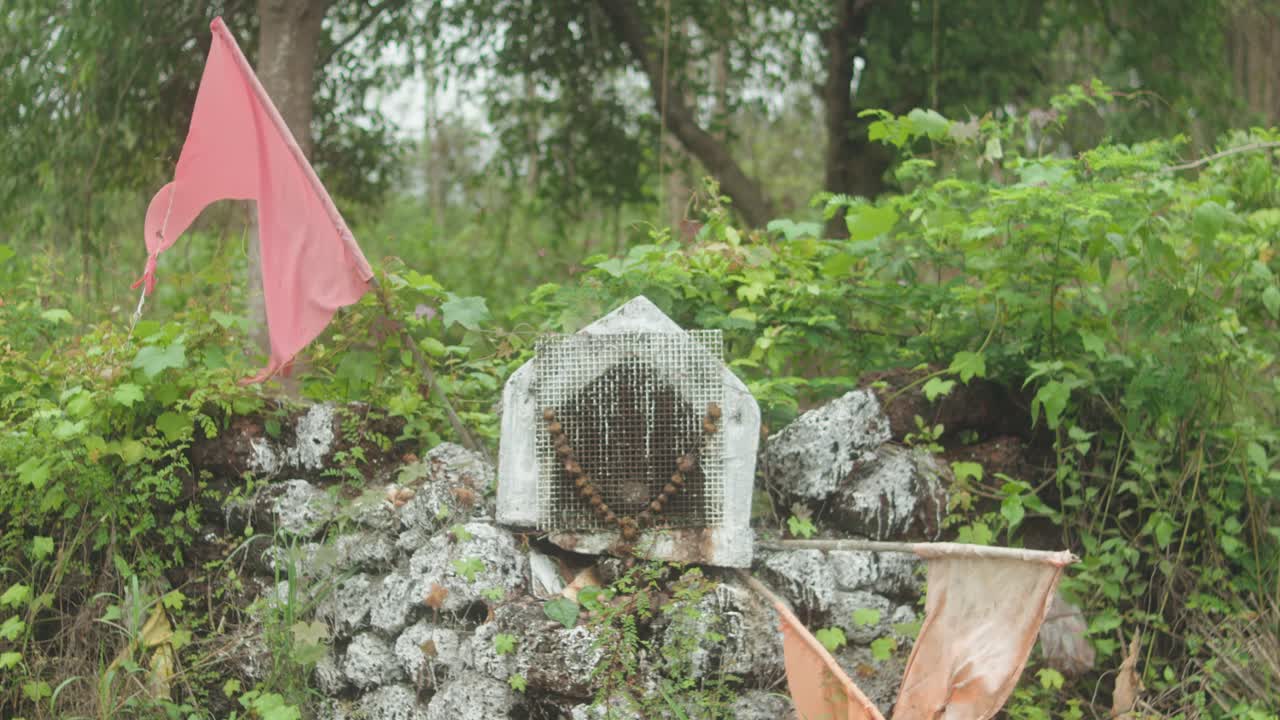Close-up of a white rural shrine adorned with prayer beads and red flag, set in natural vegetation