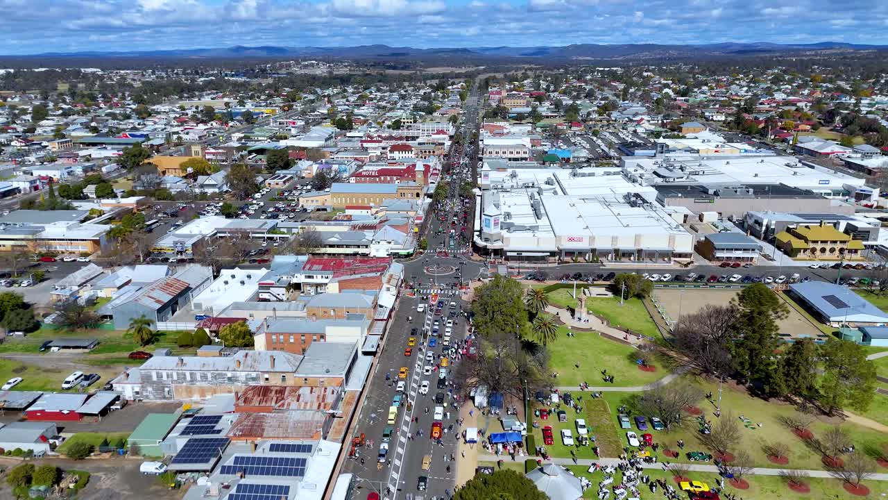 Drone captures a sunny aerial view over a busy town center, showcasing festival crowds, parked cars, and roundabouts along tree-lined streets