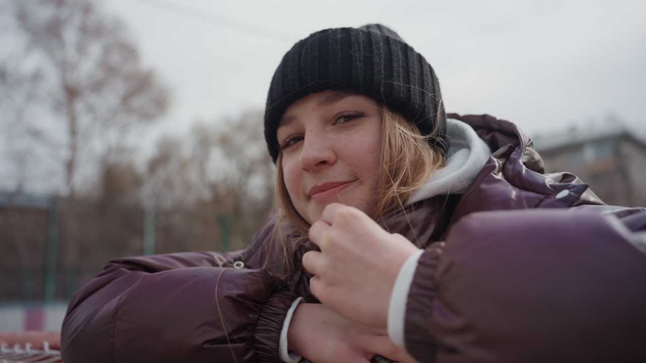 Портрет улыбающейся белой женщины, опирающейся на перила, крупный план, в черной шапке и коричневой куртке, размытый фон с осенними деревьями, мягкий свет, расслабленное задумчивое выражение лица, атмосферный портрет на открытом воздухе