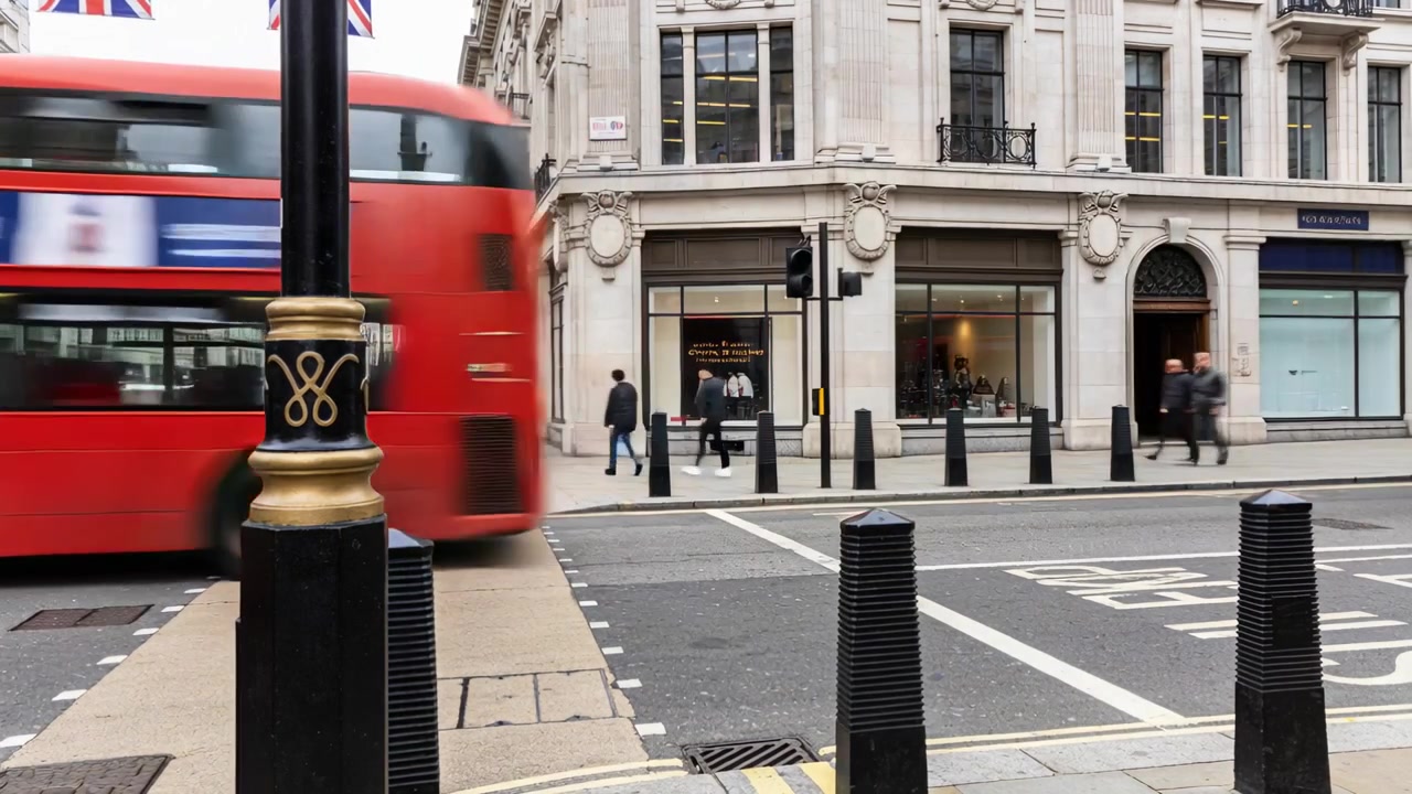 Entrando nell'inquadratura, un autobus rosso a due piani in movimento passa un attraversamento pedonale di Londra, con un lampione dorato e dissuasori.