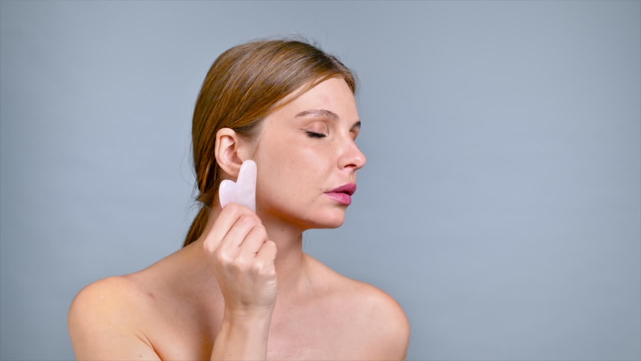 Una giovane donna caucasica si sta facendo un massaggio al viso e al collo usando una Gua Sha, guardando verso la fotocamera. Rallentatore