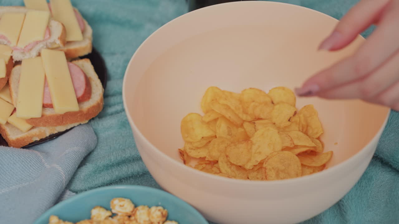Close up of hand reaching into bowl of crispy potato chips placed on soft blue fabric with cheese sausage sandwiches and caramel popcorn nearby