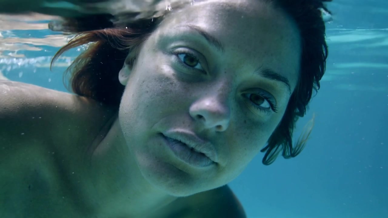 Câmera gravando, mulher prendendo a respiração e fixando o olhar na superfície da piscina, ombros à mostra, com ondulações.