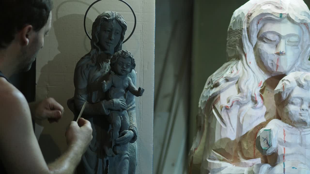 Artisan carefully perfects a clay model of Madonna and Child with a tool, while a partially carved wooden version stands nearby in a traditional studio environment