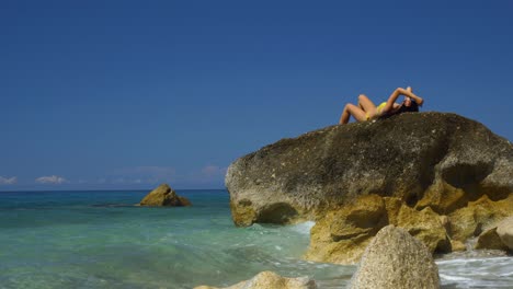 Mujer-En-Bikini-Tomando-El-Sol-Tumbada-En-Un-Acantilado-De-Granito-Bañado-Por-Agua-De-Mar-Turquesa-Cristalina-En-Una-Playa-Tranquila